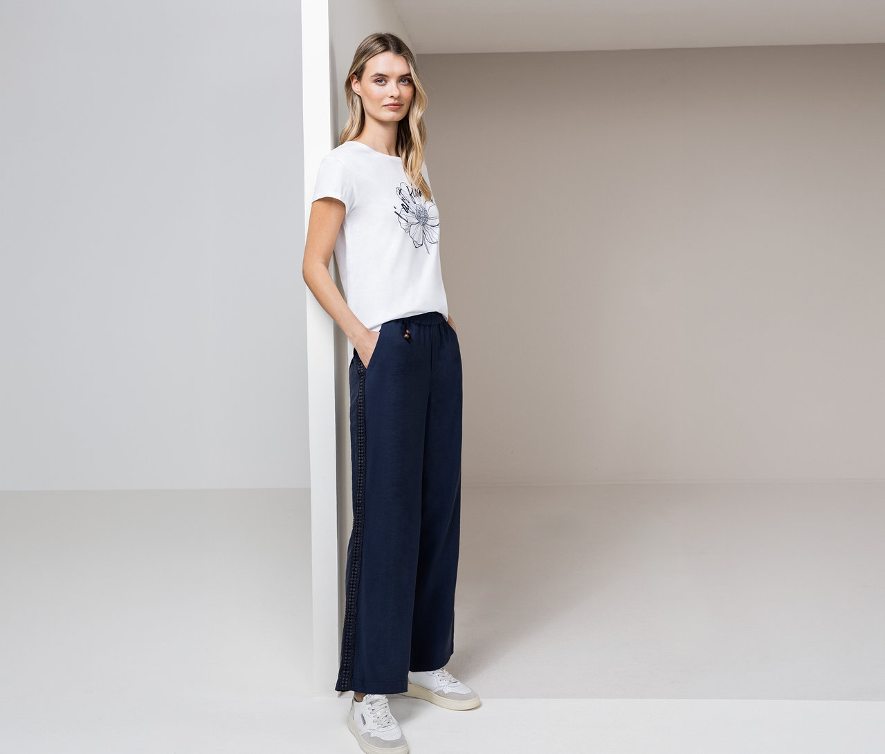 Une femme pose dans un espace lumineux, portant un t-shirt Street One, un pantalon bleu et des baskets blanches.