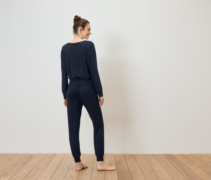 Femme avec un chignon, vue de dos, portant un haut à manches longues bleu marine et un pantalon de jogging, sur un plancher en bois devant un mur blanc.