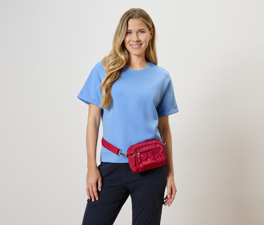 Une femme porte un t-shirt bleu clair et un sac banane rouge devant un fond neutre.