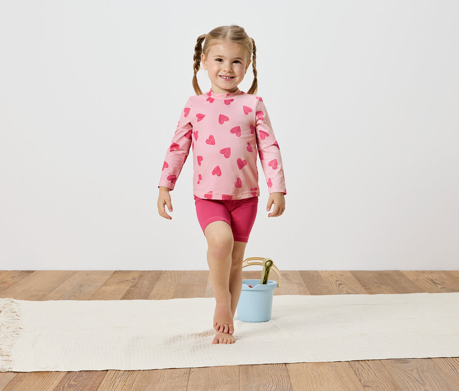 Fillette avec des tresses, portant un haut rose à cœurs et un short, en équilibre sur un tapis blanc à côté d'un seau bleu clair.