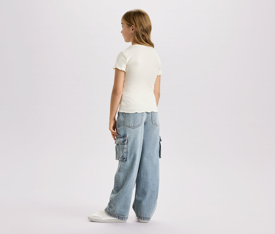 Fillette vue de dos, portant un haut court crème, un jean cargo ample et des baskets blanches sur fond blanc.