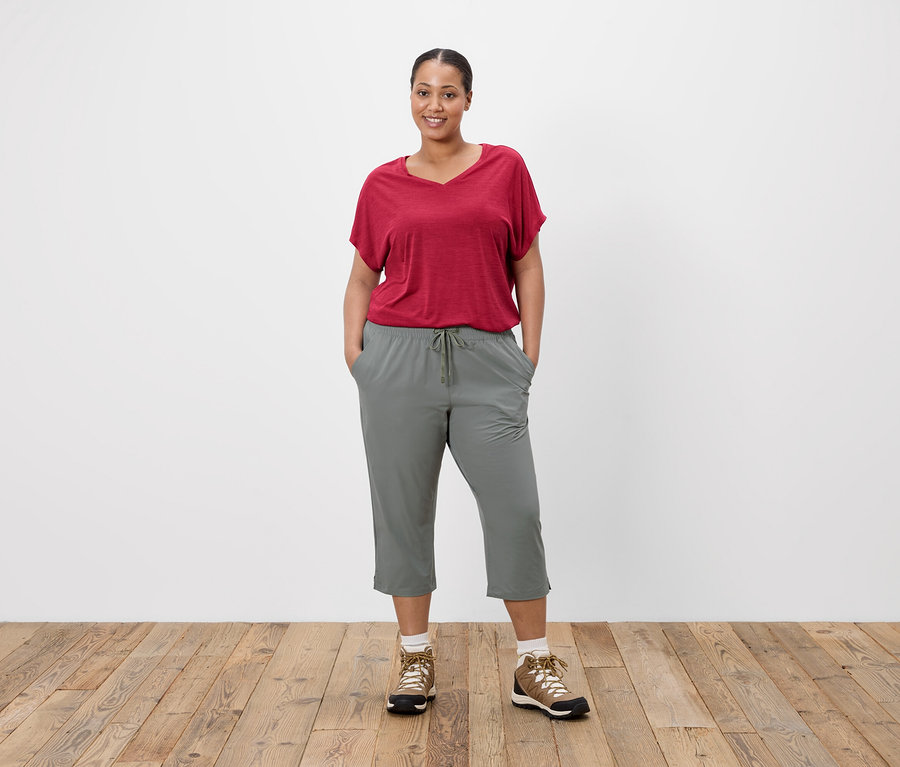 Femme en t-shirt rouge et pantalon gris debout devant un mur blanc.