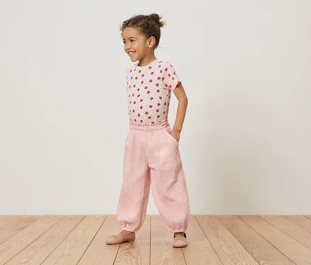 Une jeune fille se tient sur un plancher en bois, portant un t-shirt rose à fraises et un pantalon rose.