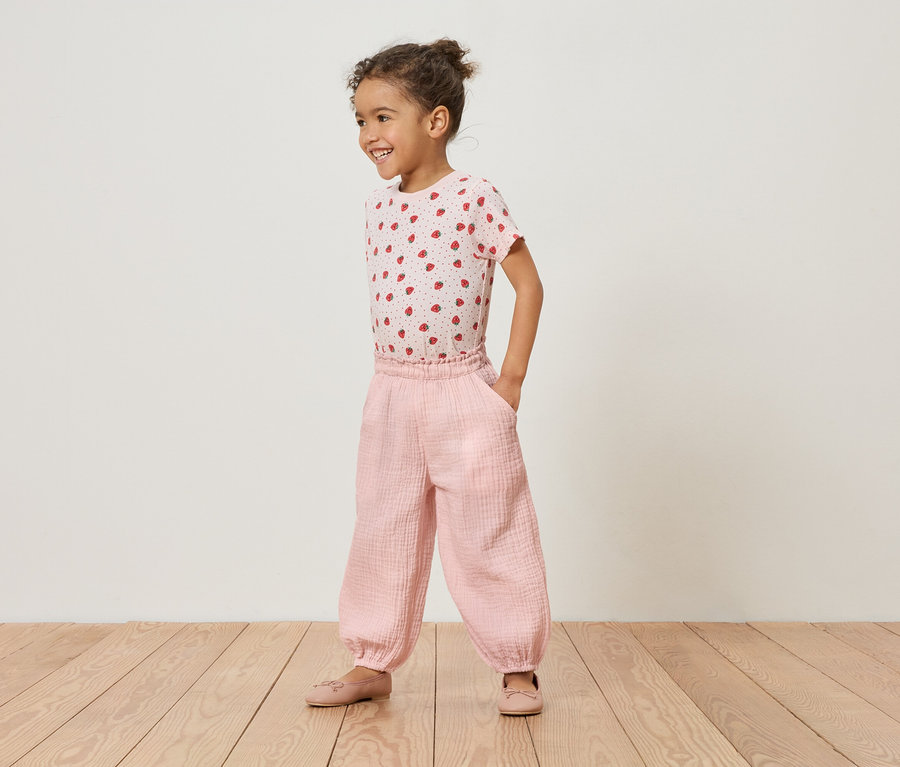 Une jeune fille se tient sur un plancher en bois, portant un t-shirt rose à fraises et un pantalon rose.