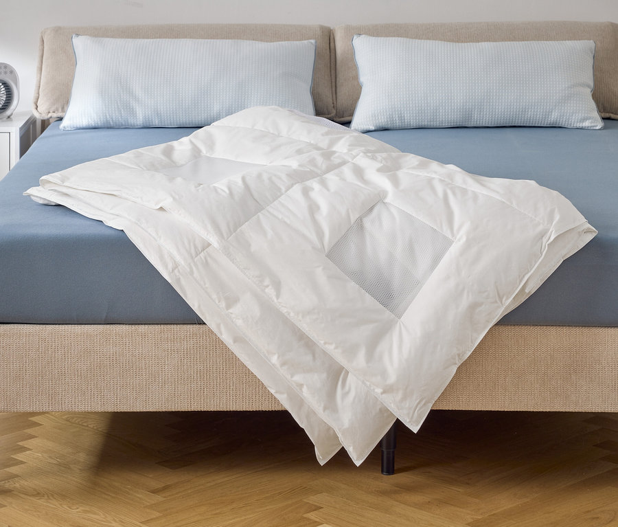 Une couette blanche est posée sur un lit avec un drap-housse bleu et deux oreillers. Un ventilateur est posé sur la table de chevet.