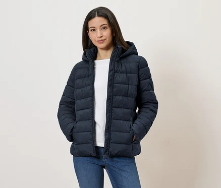 Une femme portant une doudoune à capuche bleu marine, un t-shirt blanc et un jean, se tient les mains dans les poches devant un mur blanc.