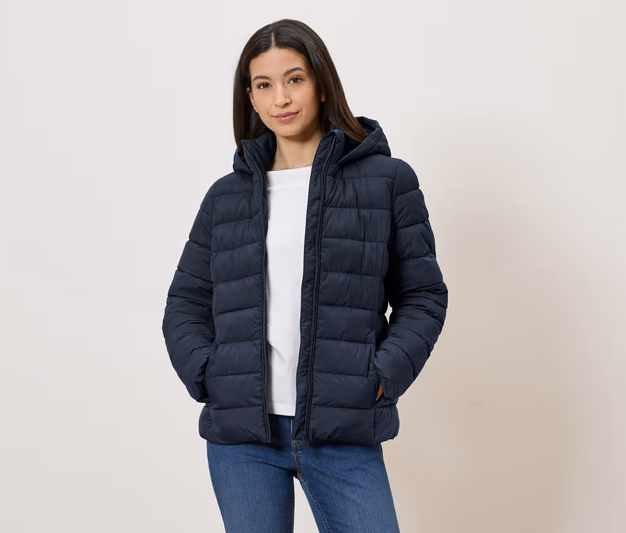 Une femme portant une doudoune à capuche bleu marine, un t-shirt blanc et un jean, se tient les mains dans les poches devant un mur blanc.