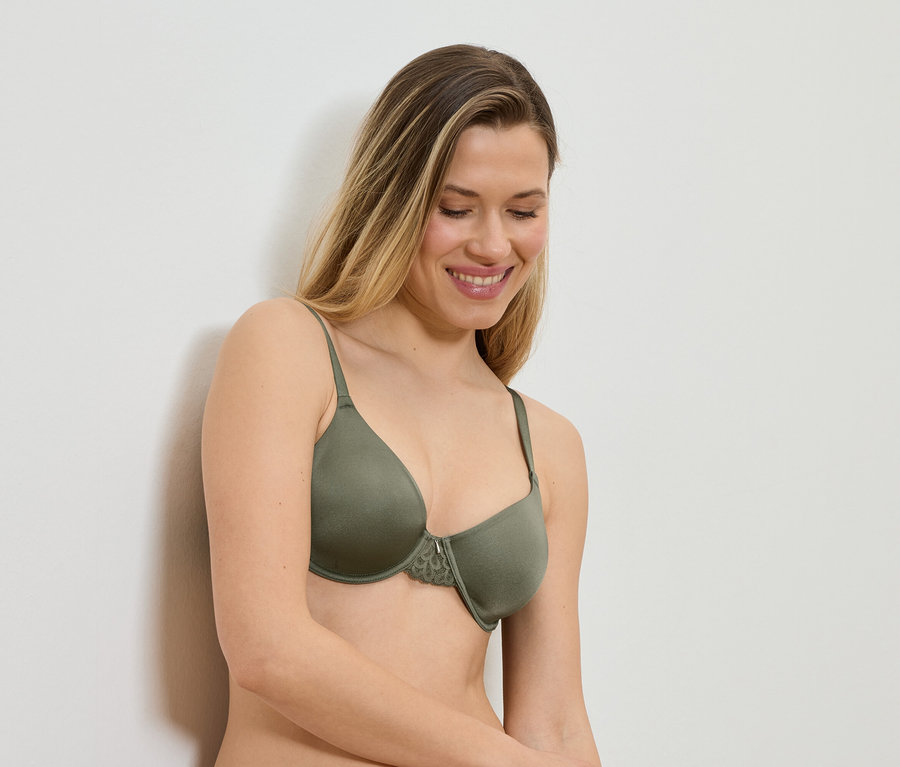Femme souriante aux longs cheveux blonds portant un soutien-gorge vert olive avec de la dentelle, appuyée contre un mur blanc.