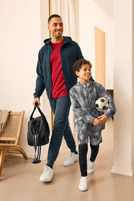 Un homme avec un sac de sport et un enfant avec un ballon de foot, tous deux portant des vêtements de sport. L'homme porte une jaquette en sweat et un pantalon en sweat. L'enfant porte une veste coupe-vent pour enfant.