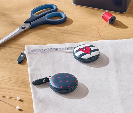 Sur une table en bois sont posés des ciseaux, une bobine de fil rouge, une boîte, deux mètres ruban et du tissu.