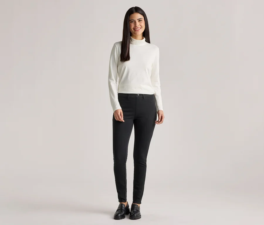 Une femme aux longs cheveux foncés porte un pull à col roulé blanc, un pantalon noir et des chaussures noires.