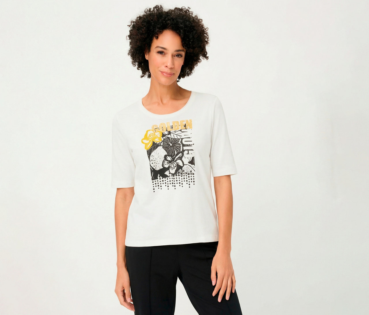 Une femme porte un T-shirt 3/4 Olsen blanc avec un motif floral et un pantalon noir.