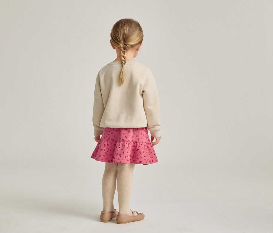 Vue de dos d'une fillette avec une tresse blonde portant un pull beige, une jupe rose à volants et des ballerines.