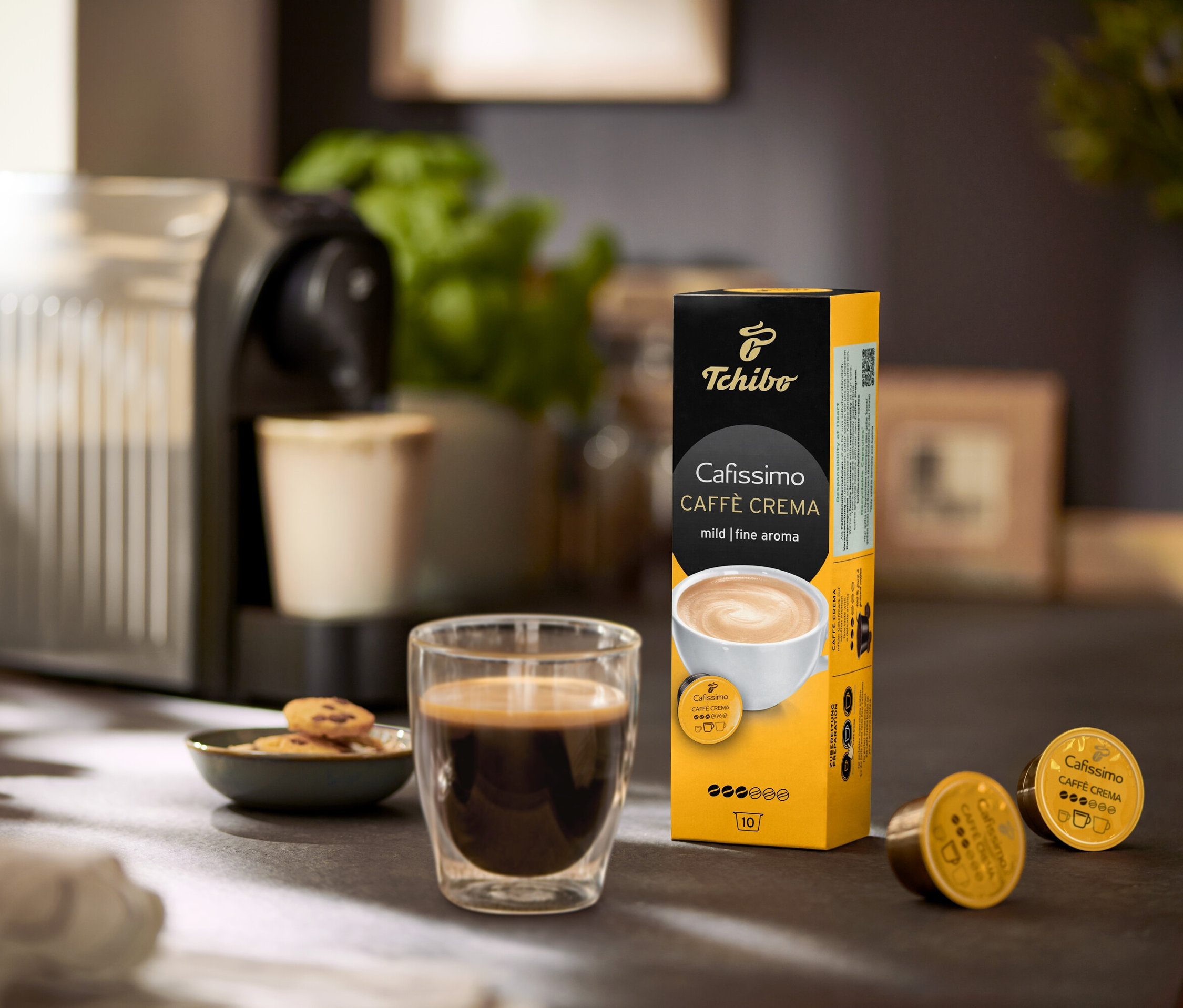 Sur une table, il y a une machine à café, un verre de café et un paquet de Cafissimo Caffè Crema mild (doux) – 120 capsules.