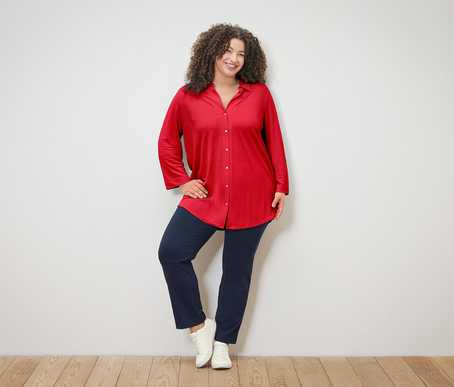 Une femme aux cheveux bouclés porte un chemisier rouge, un pantalon bleu et des baskets blanches, et pose devant un mur blanc.