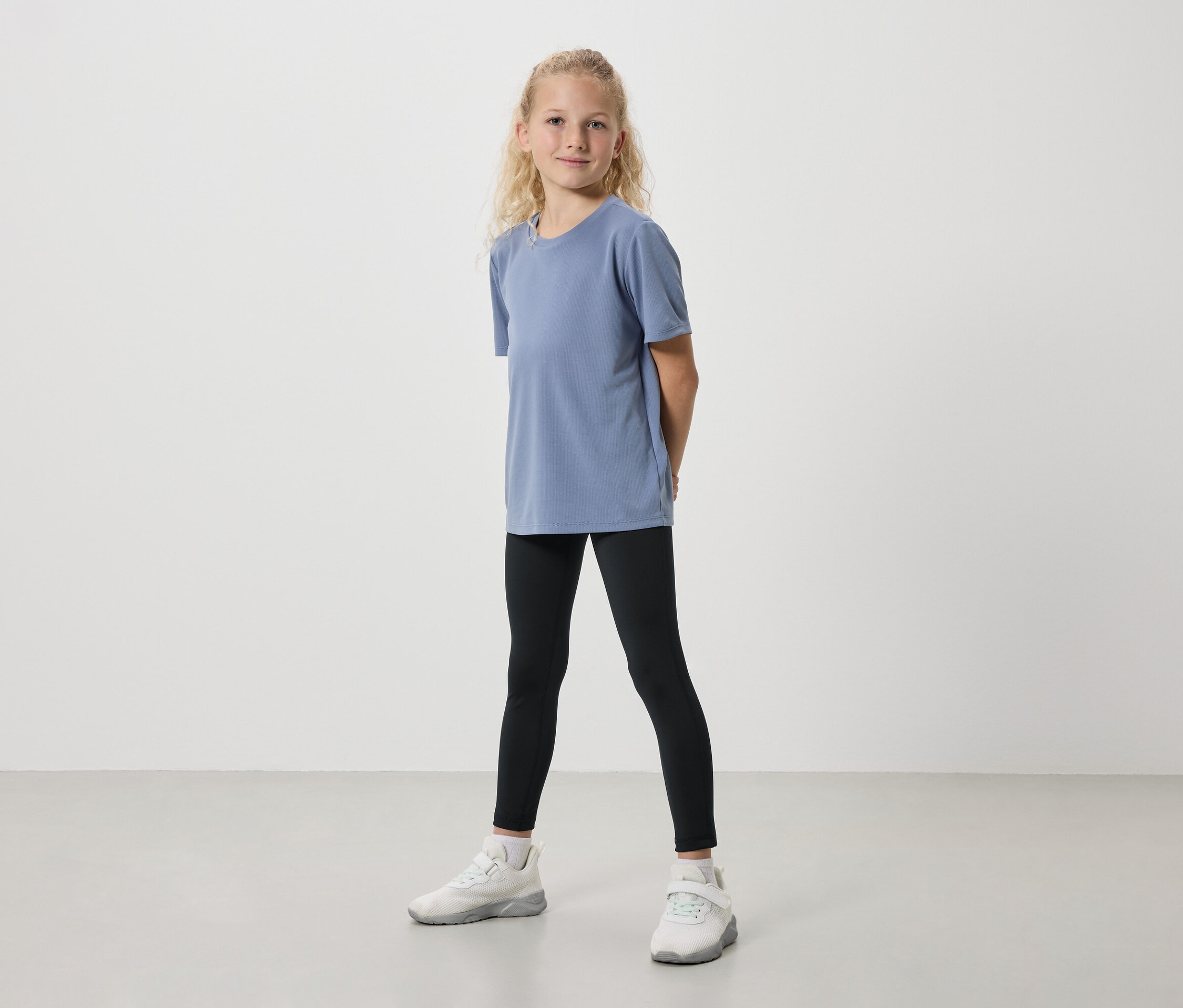 Une fille se tient de face avec un t-shirt bleu, un legging noir et des baskets blanches sur un fond clair.