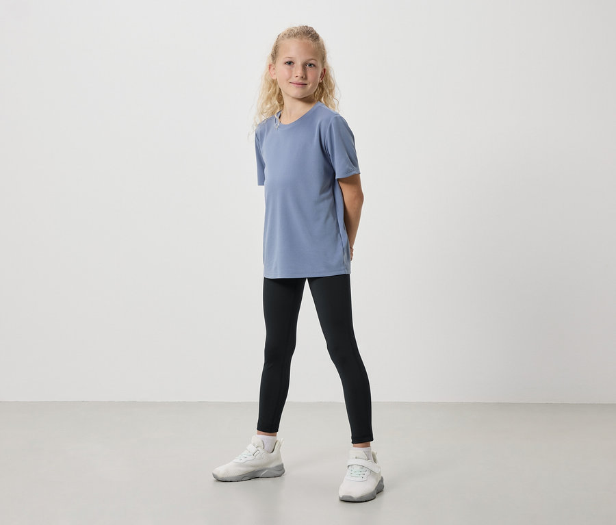 Une fille se tient de face avec un t-shirt bleu, un legging noir et des baskets blanches sur un fond clair.