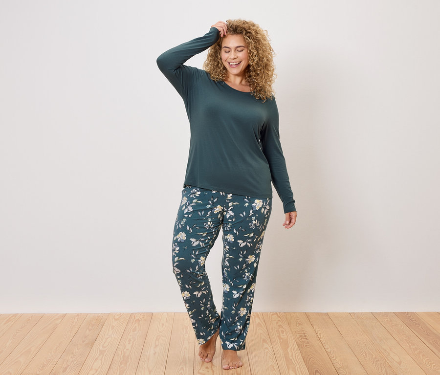Une femme aux cheveux bouclés porte un haut à manches longues vert foncé et un pantalon de pyjama vert foncé à motifs floraux.