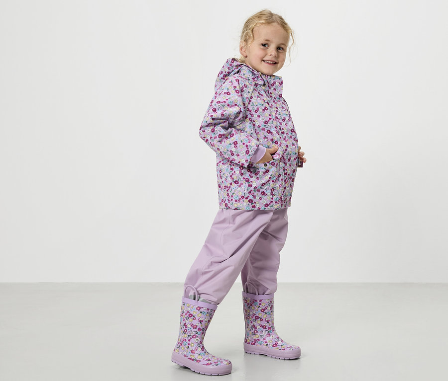 Une fillette blonde porte un manteau de pluie violet à motifs floraux, un pantalon de pluie violet et des bottes en caoutchouc assorties.