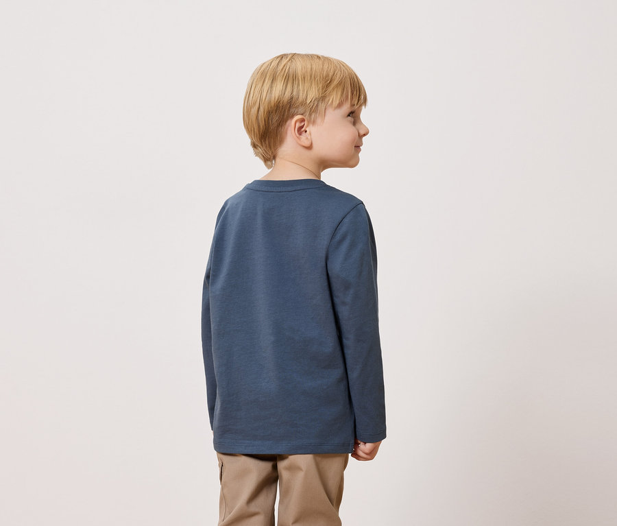 Un garçon blond se tient dos à la caméra, portant un t-shirt bleu à manches longues et un pantalon kaki.