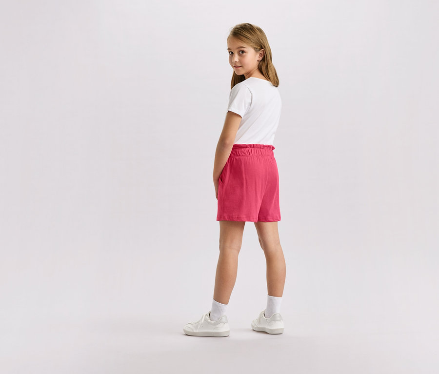 Fillette blonde portant un t-shirt blanc, un short rose et des baskets blanches, regardant la caméra.