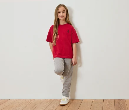 Fillette aux longs cheveux bruns, portant un t-shirt rouge et un jogging gris, appuyée contre un mur blanc.