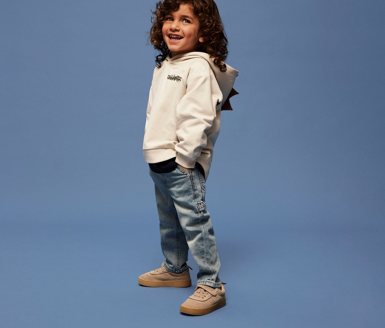 Un garçon aux cheveux bouclés porte un sweat-shirt avec application dino en 3D pour enfant NAME IT, un jean et des baskets sur fond bleu.