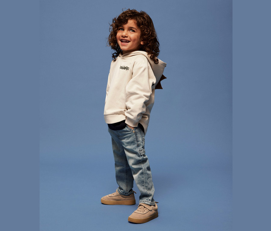 Un garçon souriant aux cheveux bouclés se tient sur un fond bleu en sweat à capuche beige, jean bleu clair et chaussures marron.