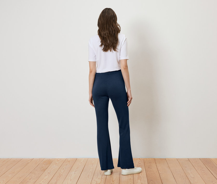 Vue de dos d'une femme aux longs cheveux bruns portant un t-shirt blanc, un pantalon bootcut bleu marine et des baskets blanches sur un parquet devant un mur couleur crème.