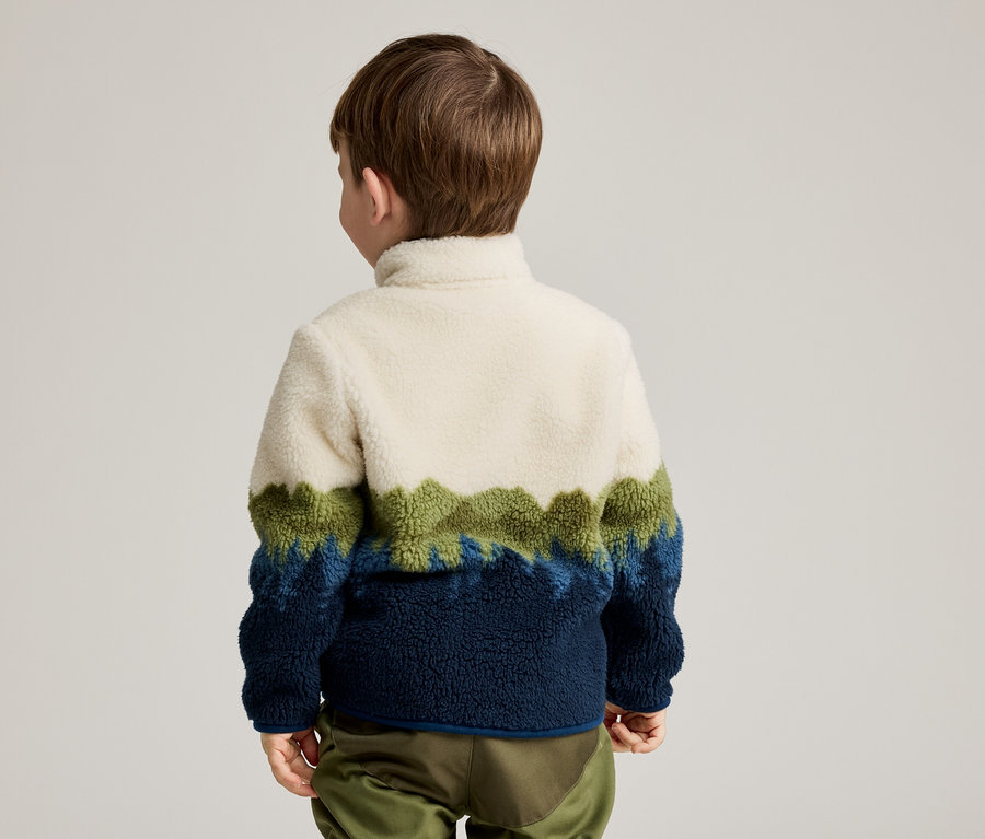 Vue arrière d'un enfant aux cheveux bruns portant un pull en polaire blanc, vert et bleu et un pantalon vert.