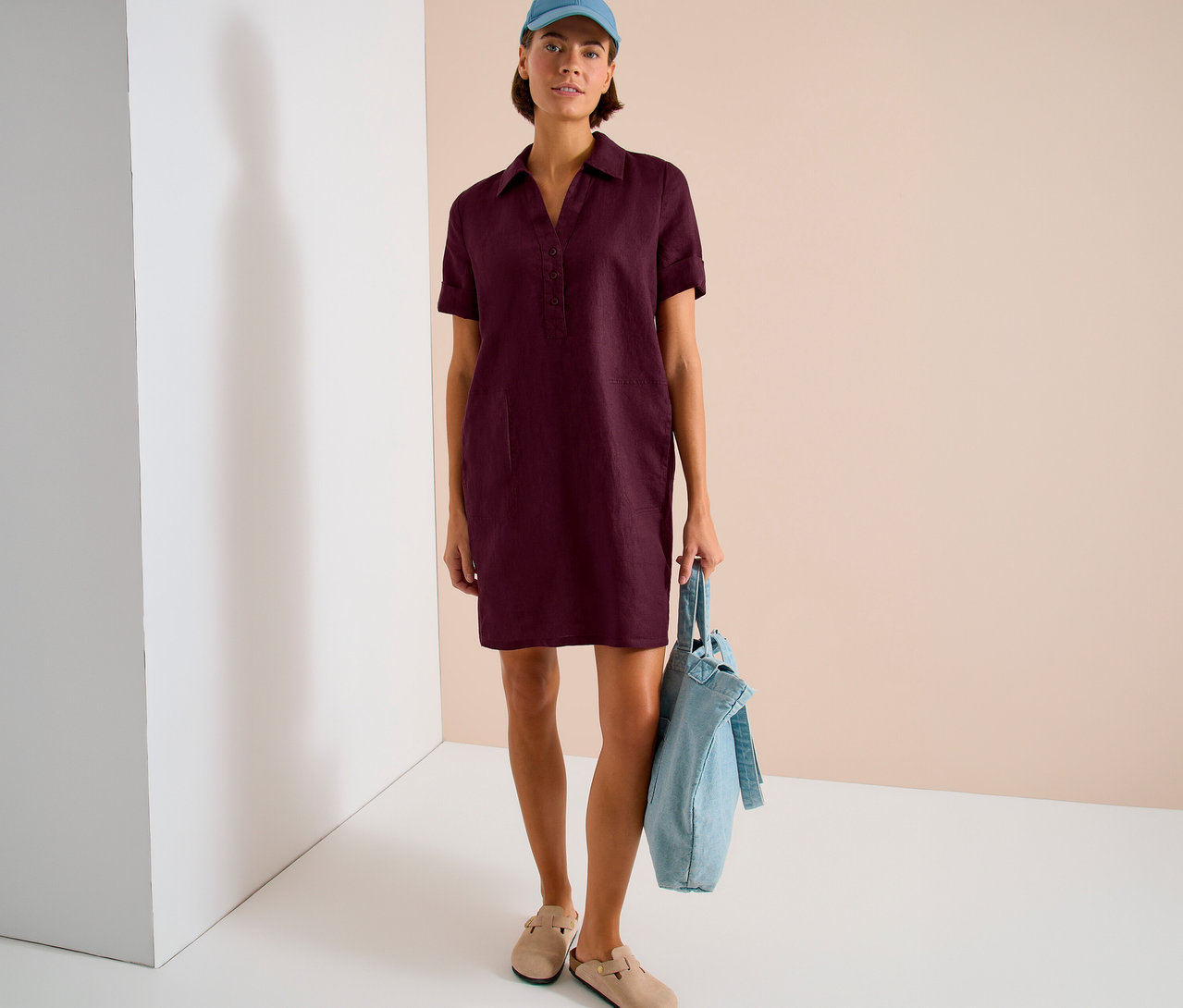 Une femme en robe chemise bordeaux, casquette bleue et sabots beiges tient un sac en tissu bleu clair.