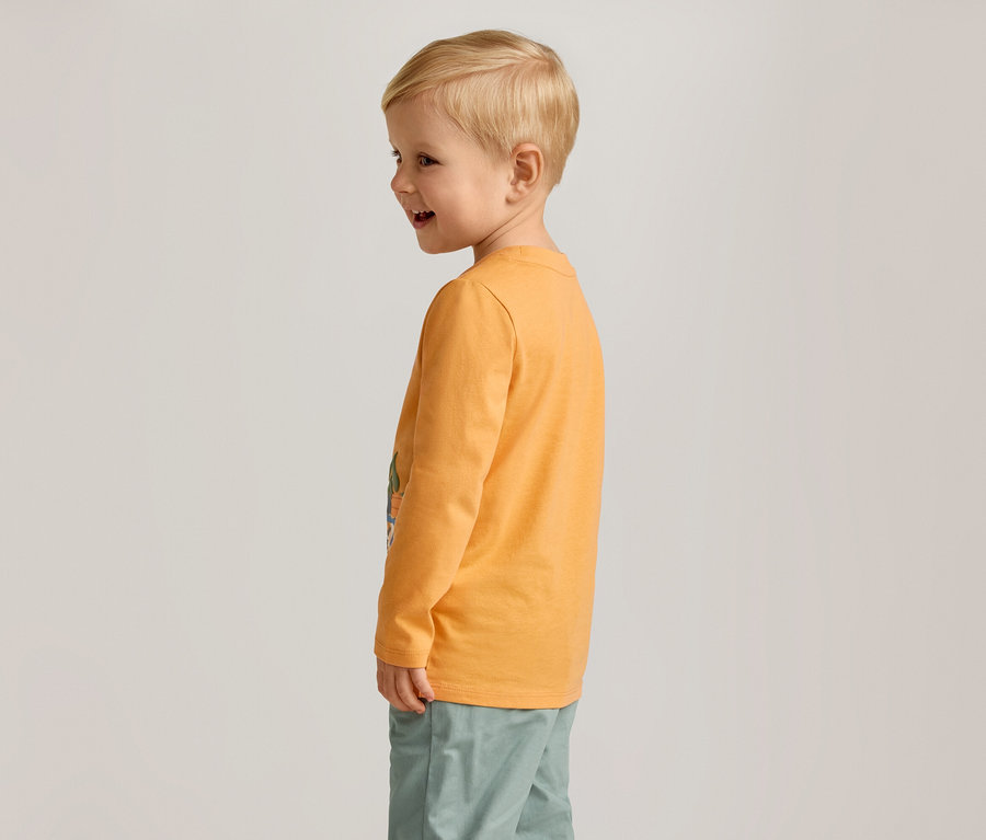 Un garçon blond souriant, vu de profil, porte un t-shirt orange à manches longues et un pantalon vert.