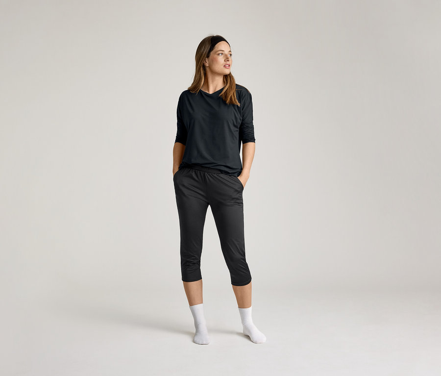 Femme portant un t-shirt de sport 3/4 noir et un pantalon foncé, mains dans les poches, chaussettes blanches, sur fond clair.