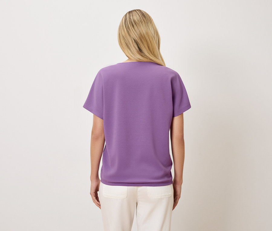 Vue arrière d'une femme aux cheveux blonds portant un t-shirt violet et un pantalon crème.