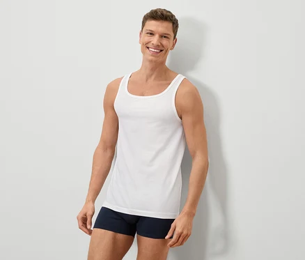 Un homme souriant aux cheveux bruns courts porte un débardeur blanc et un caleçon bleu devant un mur blanc.
