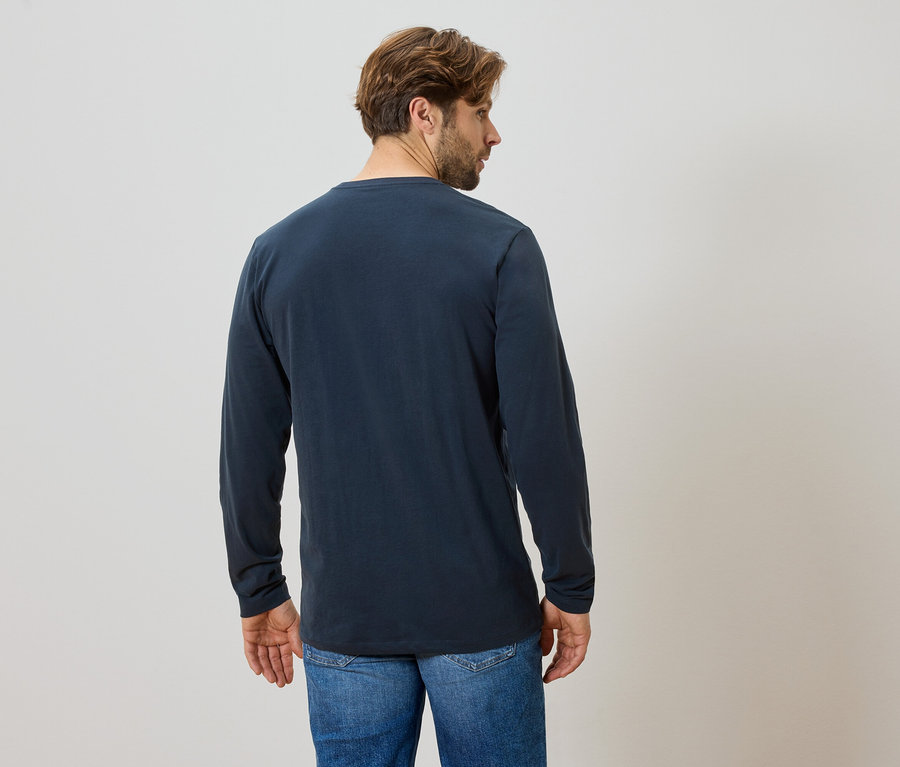 Homme vu de dos portant un t-shirt bleu à manches longues et un jean sur un fond clair.