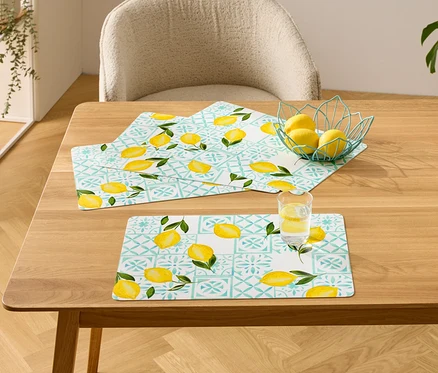 Sur une table en bois, trois sets de table à motif citron, un bol de citrons et un verre d'eau avec du citron.