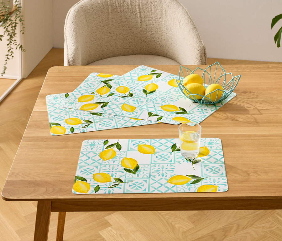 Sur une table en bois, trois sets de table à motif citron, un bol de citrons et un verre d'eau avec du citron.