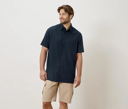 Un homme barbu porte une chemise à manches courtes bleu marine et un short cargo kaki sur un fond crème.