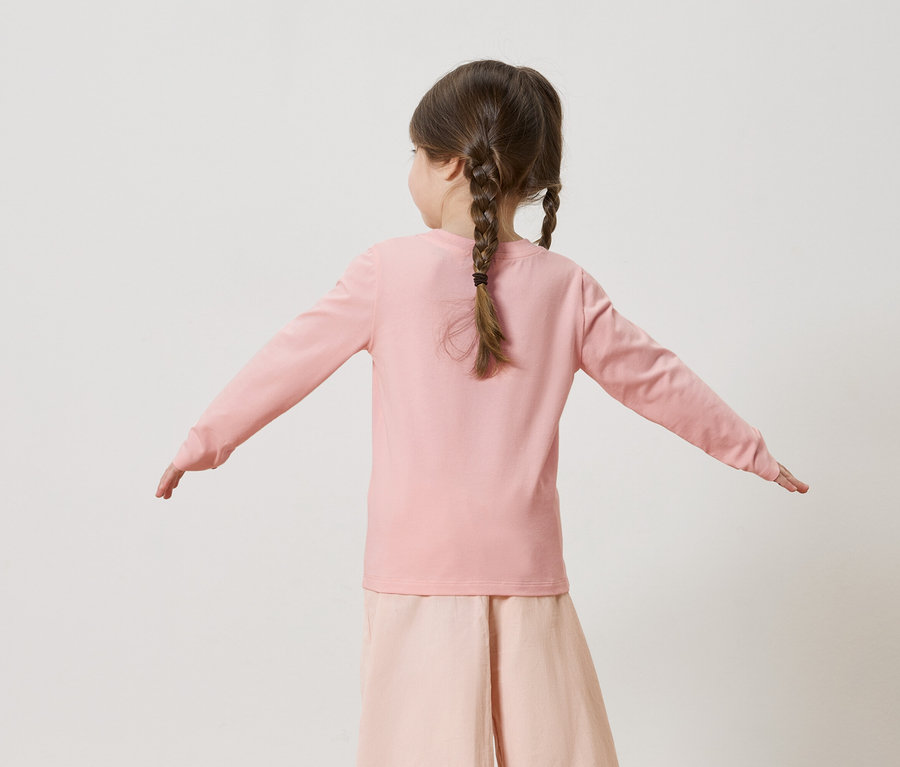 Vue de dos d'une petite fille avec des tresses portant un t-shirt rose à manches longues et un pantalon rose, les bras tendus sur les côtés.