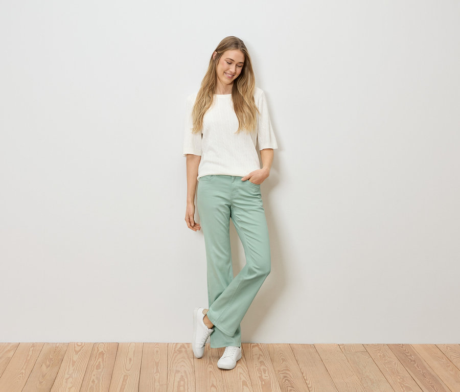 Une femme blonde portant un haut blanc, un pantalon vert et des baskets blanches se tient devant un mur blanc sur un plancher en bois.