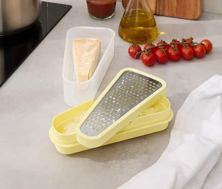 Sur un comptoir de cuisine se trouvent une casserole, des tomates en grappe, une bouteille d'huile, un bol de sauce, un récipient avec du fromage et une râpe jaune.
