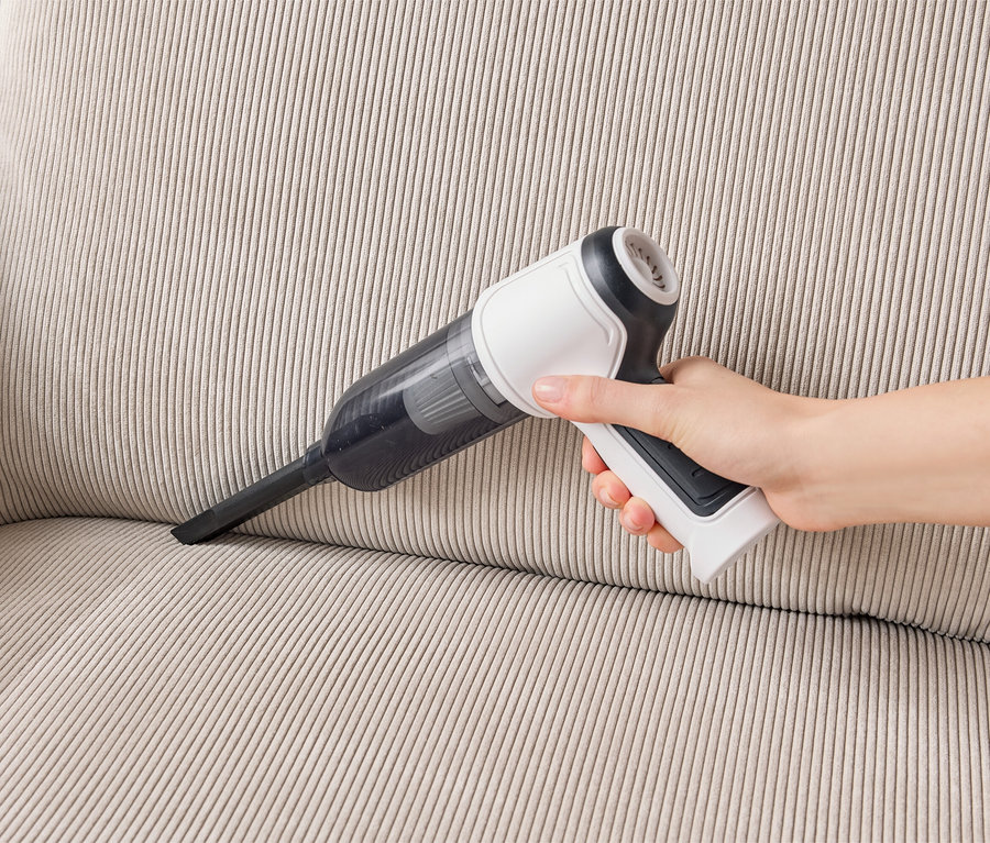Une main tient un aspirateur à main blanc et noir, aspirant un canapé beige.