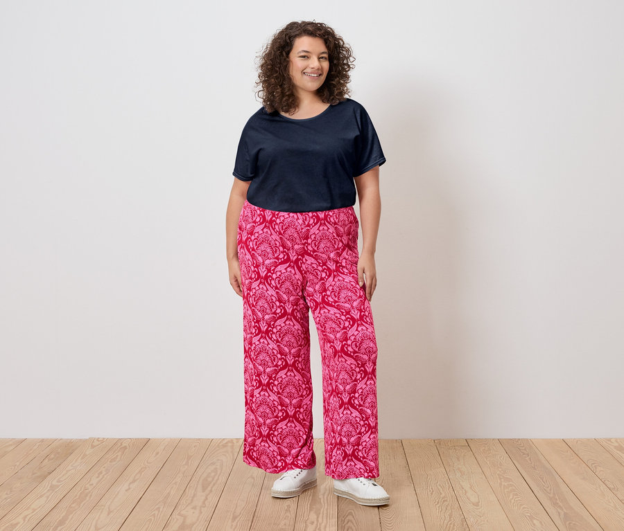 Une femme aux cheveux bouclés porte un t-shirt bleu marine, un pantalon rose à motifs et des baskets blanches.