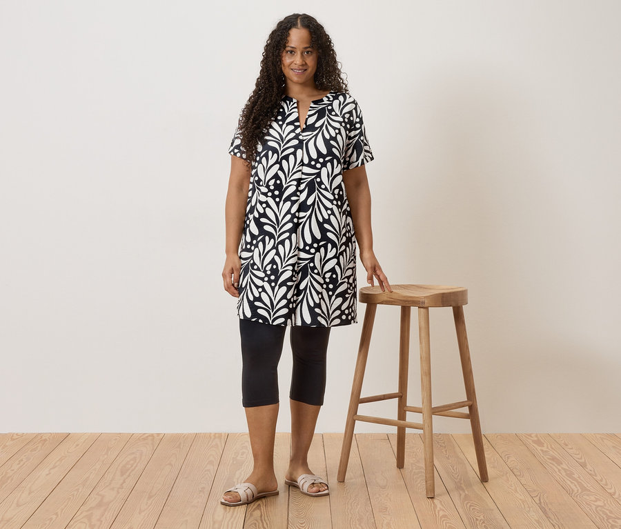 Une femme aux cheveux bouclés portant une robe noire et blanche, des leggings et des sandales se tient à côté d'un tabouret.