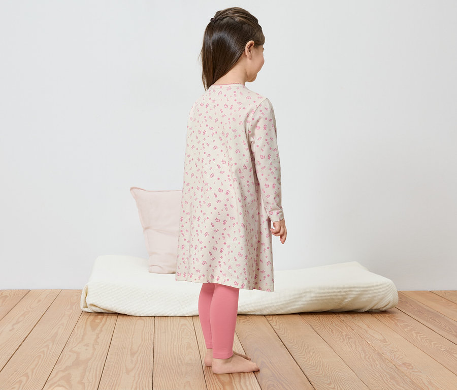 Une fille avec une tresse se tient sur un plancher en bois, portant une robe à fleurs et des leggings roses.