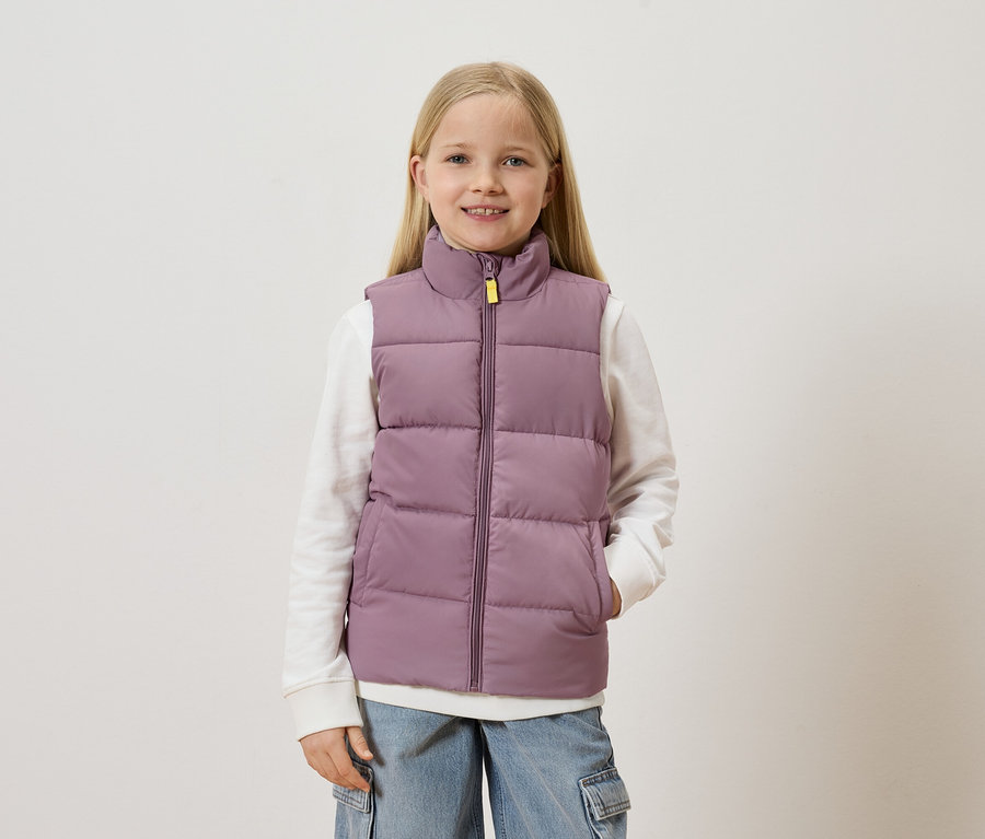 Une fille blonde porte un gilet violet sur un pull blanc et un jean bleu avec des poches.