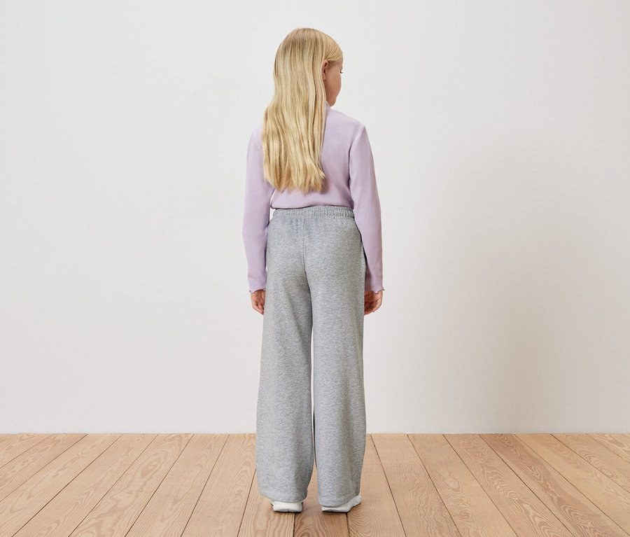 Vue de dos d'une fille blonde portant un pull lilas clair et un pantalon gris large devant un mur blanc.