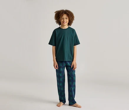 Un garçon souriant aux cheveux bouclés porte un pyjama vert foncé avec un pantalon à carreaux.