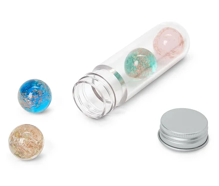 Quatre sphères pailletées bleues, turquoise, roses et beiges, dont deux à l'intérieur d'un tube en plastique transparent ouvert et deux à l'extérieur. Un capuchon argenté est posé à côté du tube.
